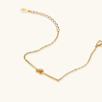 Delicate gold chain bracelet with minimalist bar and centered knot detail on cream background